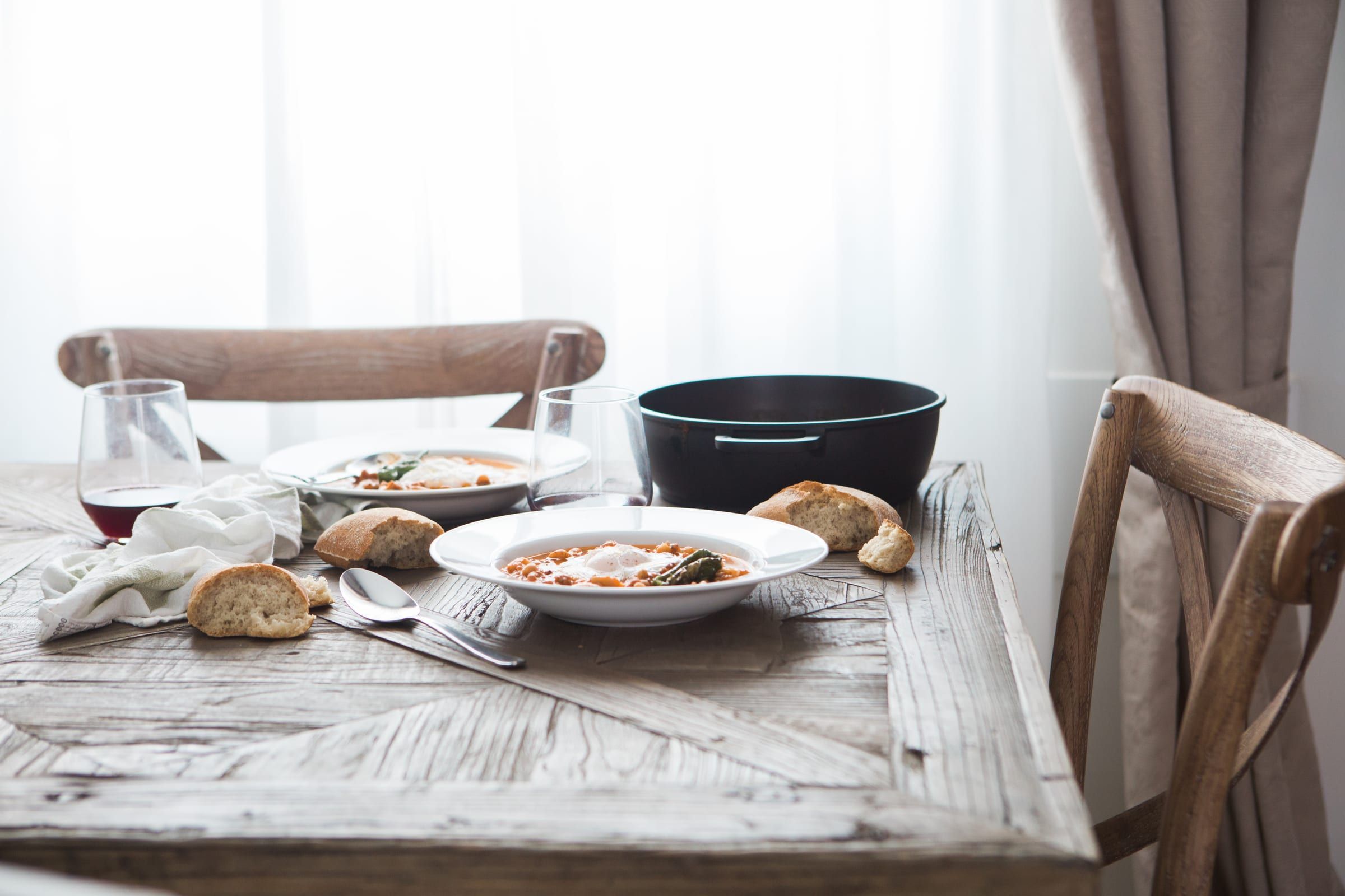 Bowls of bean stew with poached eggs, torn bread and red wine on a wooden table by a bright window