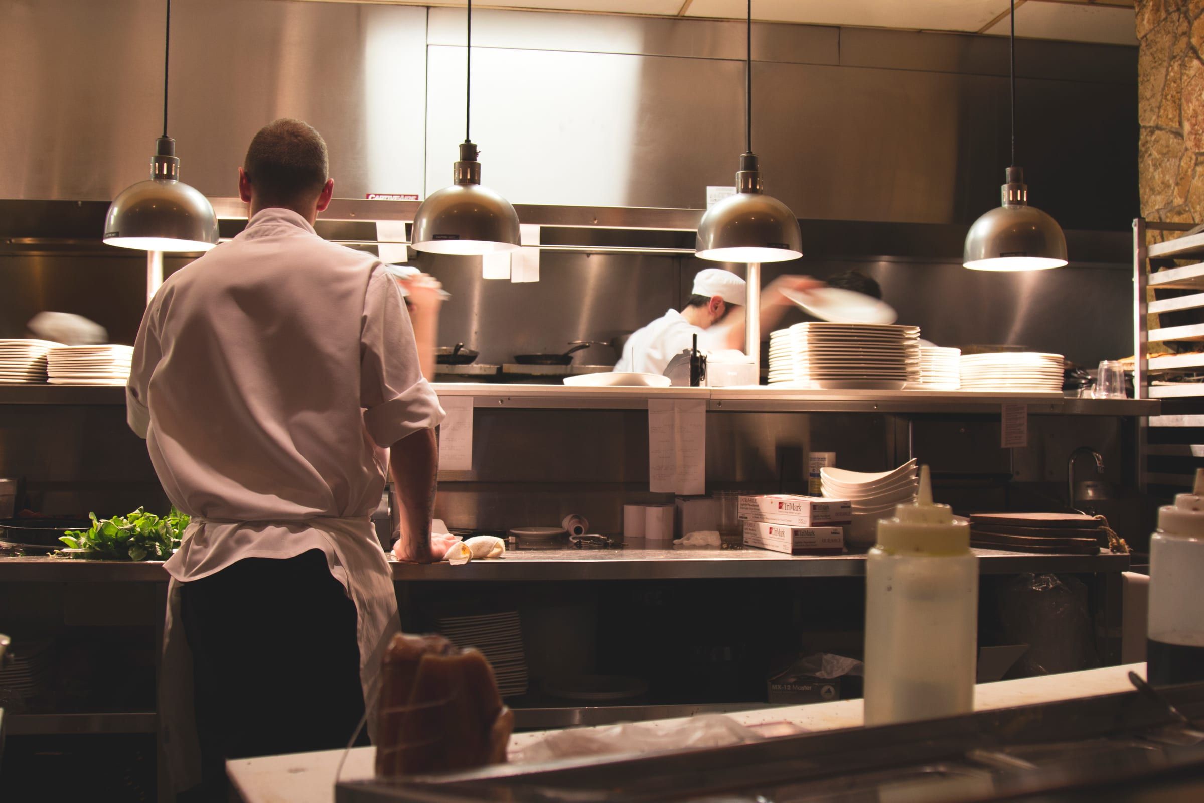 A cook at the pass in the open kitchen, under hanging lamps