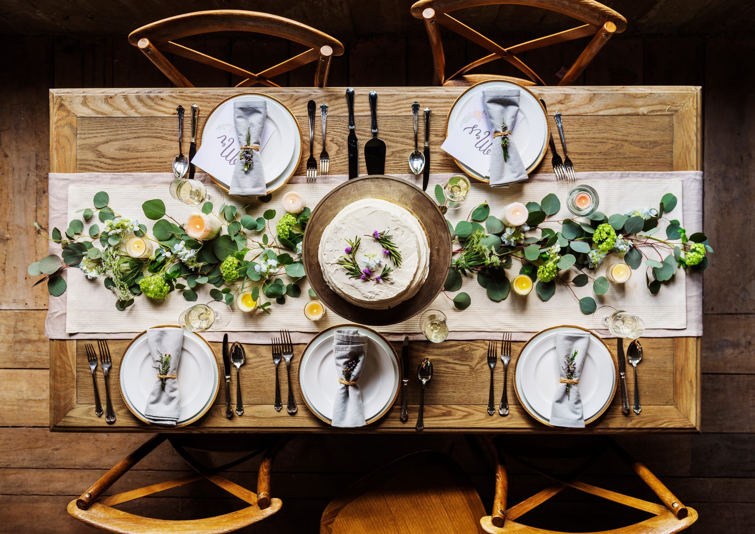 A long table seen from above, set with white plates, linen napkins and a garland of greenery